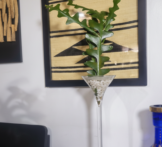 Tall glass vase with decorative plant on a surface with a blue and gold vase in the background.
