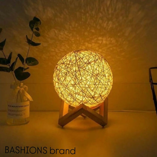 Woven ball lamp on a stand with a plant and bottle in the background, illuminated by the lamp.